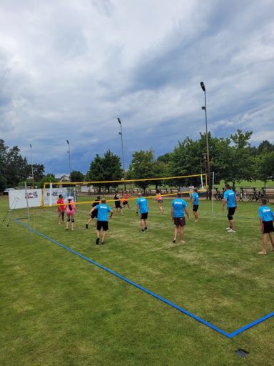 Gruppe von Menschen spielt Volleyball auf einer Wiese unter bewölktem Himmel.
